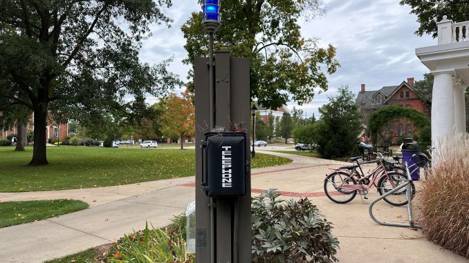 A telephone covered by a black case with a blue light on top of it stands outside of Vulgamore Hall, surrounded by trees on the left and bicycles on the right.