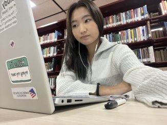 A young woman dressed in a gray and white sweater sits at a table in front of a laptop with earphones in her ears. She gazes thoughtfully to the side, and a pen and notebook rest on the table nearby.