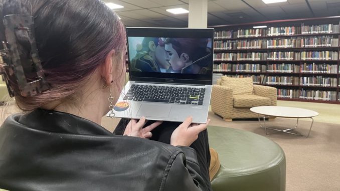 A close-up photo taken over the shoulder of a young woman watching a TV show on a laptop. On-screen, two animated characters kiss. In the background of the photo are furniture and bookshelves.