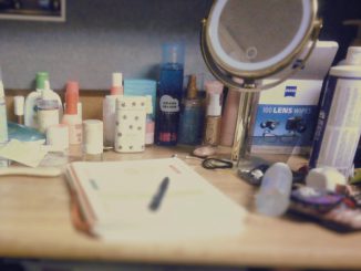 A desk littered with beauty products and a mirror. There is a journal with a pen in the middle, and the photo is a bit grainy.