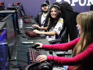 A young woman in a long-sleeve pink shirt plays a video game on a gaming computer while looking towards and speaking to another young woman who is smiling. The two are sitting in a row of gaming computers, with another student farther down the row looking in their direction.