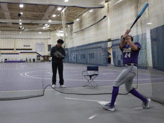 A baseball player wearing a purple and gray uniform swings a bat in an indoor batting cage, standing outside the batting cage a young man observes and takes notes.