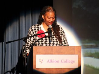 A woman in a black and white patterned shirt with a red rose smiles as she stands behind a podium that reads “Albion College.” A microphone is positioned in front of her and a spotlight illuminates a circle behind her.