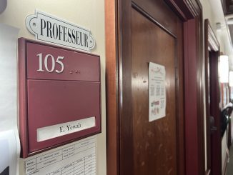 In the foreground, a red plaque with the title “professeur” above it and “E. Yewah” written within it. Next to the plaque is a wooden door with a course offerings paper attached to it.