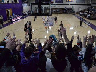 A packed student section raises their hands as a basketball player prepares to take a free throw in a college gym. The court has purple and gold branding, with players and referees positioned for the shot.