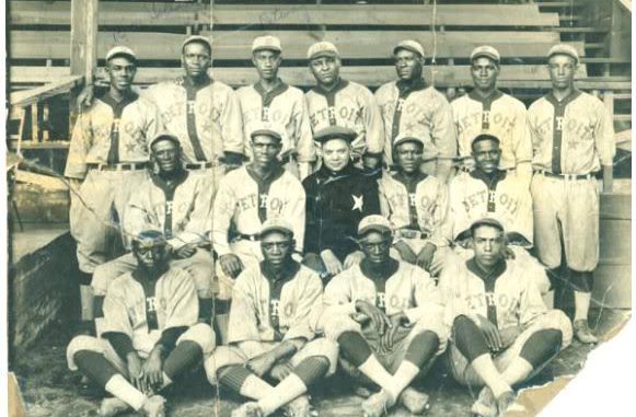 An aged black-and-white photograph shows players on an African-American baseball team organized into three rows and pose for the camera. The players are wearing baseball uniforms that say Detroit on the front, while the owner, who is seated in the center, is wearing a dark coat with a star on the chest.