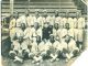 An aged black-and-white photograph shows players on an African-American baseball team organized into three rows and pose for the camera. The players are wearing baseball uniforms that say Detroit on the front, while the owner, who is seated in the center, is wearing a dark coat with a star on the chest.