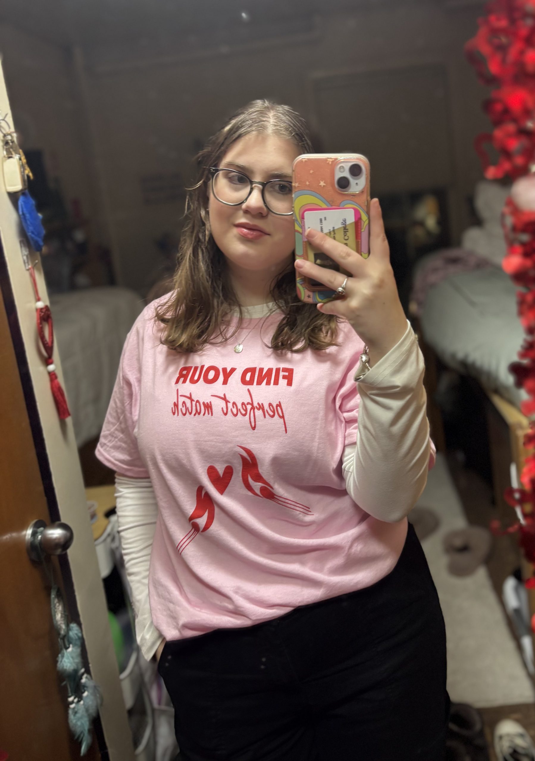 A woman with brown hair and green glasses poses in a mirror holding a phone with a pink phone case. The woman is wearing a baby pink t-shirt that reads “Find your perfect match” with a white shirt underneath and black jeans. 