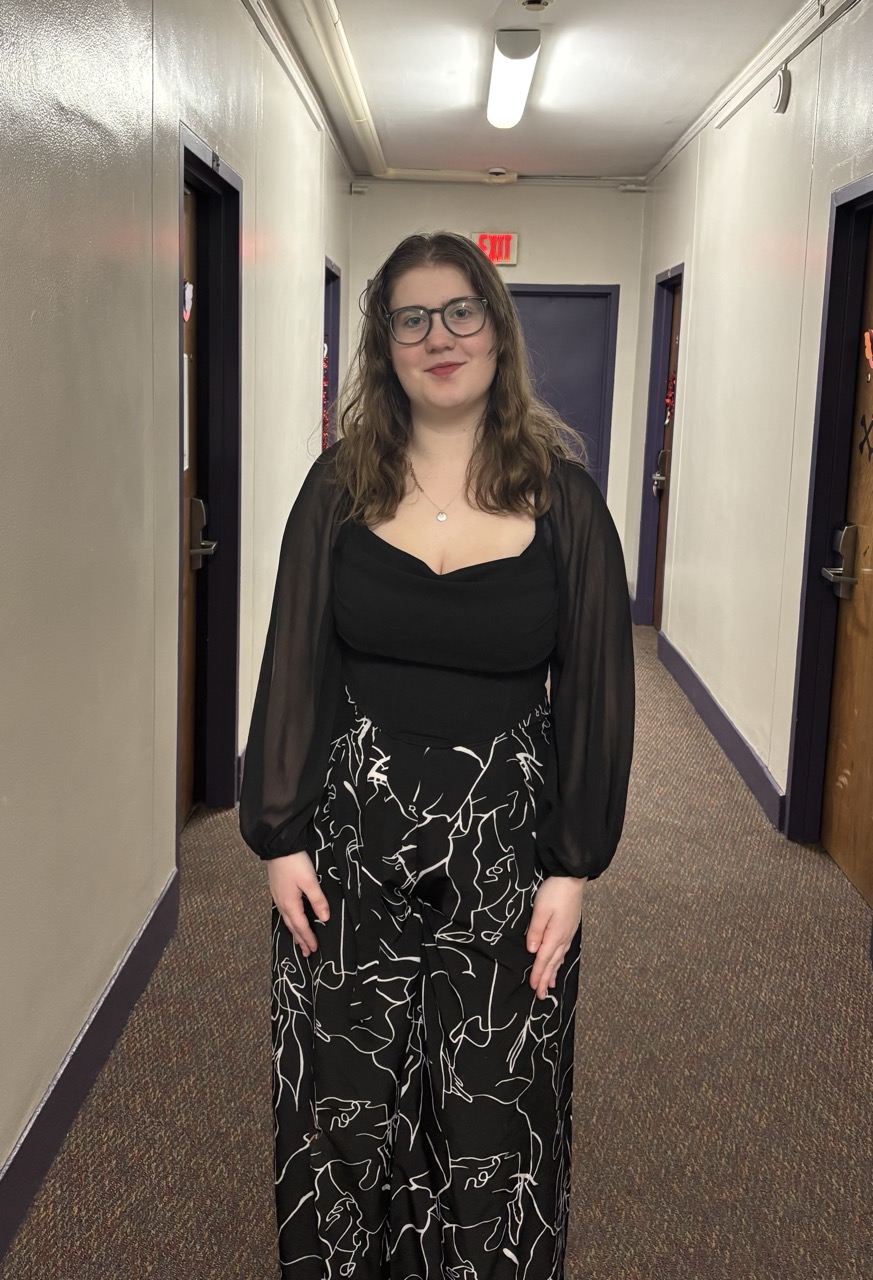 Woman with brown hair and green glasses stands in the middle of a hallway with purple trim and wooden doors. The woman is wearing a black blouse with sheer sleeves and black pants with white lines in a random pattern. 