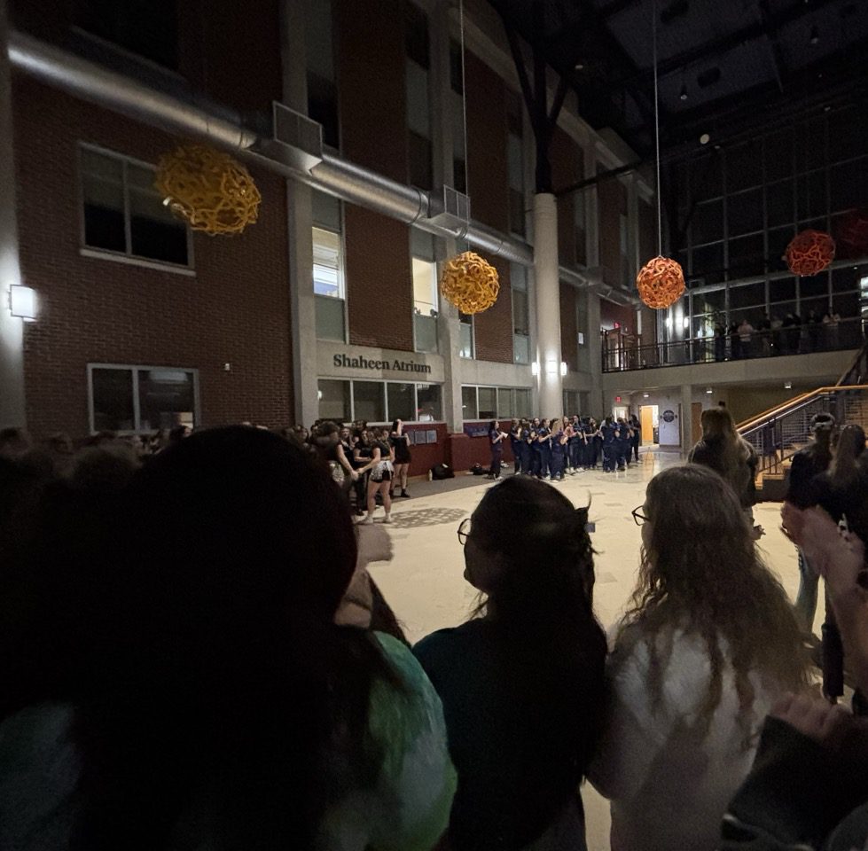 A wide open room with large yellow and orange ball sculptures hanging from the ceiling. In the foreground there are the backs of four women’s heads and one woman wearing bear ears clapping. In the background are three groups of women gathered together cheering. 