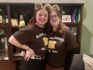 Two young women stand wearing matching dark-brown t-shirts that read“Kappa Delta Est. 1897” with fluffy brown bear ears. The woman on the left wears black jeans with a silver belt, and the woman on the right wears brown ripped jeans.