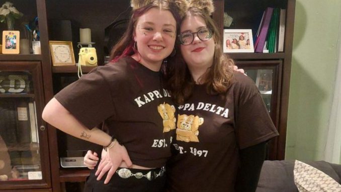 Two young women stand wearing matching dark-brown t-shirts that read“Kappa Delta Est. 1897” with fluffy brown bear ears. The woman on the left wears black jeans with a silver belt, and the woman on the right wears brown ripped jeans.