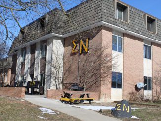 A largely brick building with the Greek letters Sigma Nu written on benches outside the door in yellow paint, displayed as a yellow sign on the side of the house and painted in yellow paint on a rock to the right of the house.