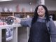 A young adult standing amidst a ceramics studio smiles as they look at the camera. The individual is wearing a gray sweatshirt with a black apron over it, and they have a hand on a ceramic sphere with multiple holes in it.