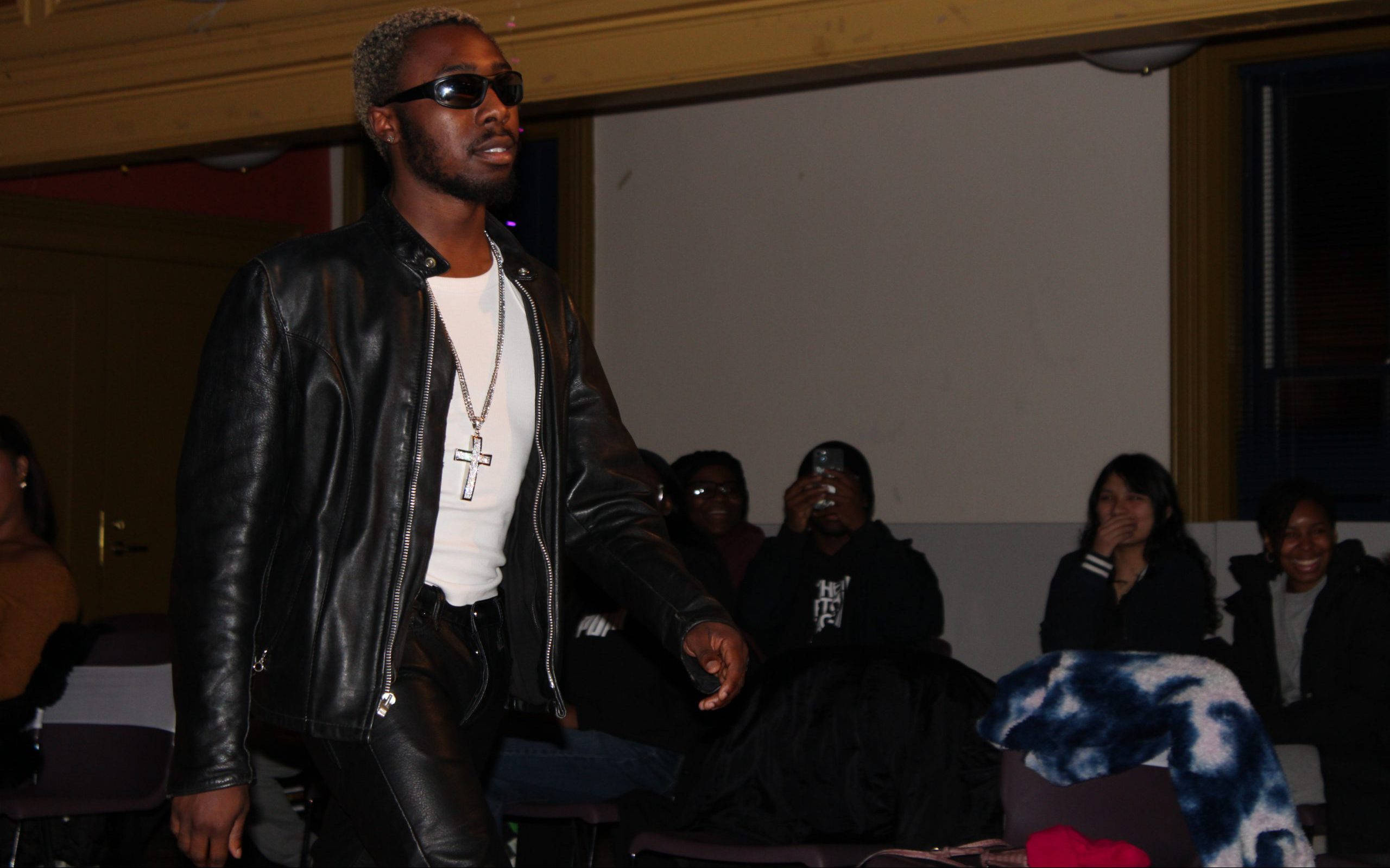 A young man wearing a white undershirt with a cross necklace along with black leather pants and jacket walks in a room with a seated audience.