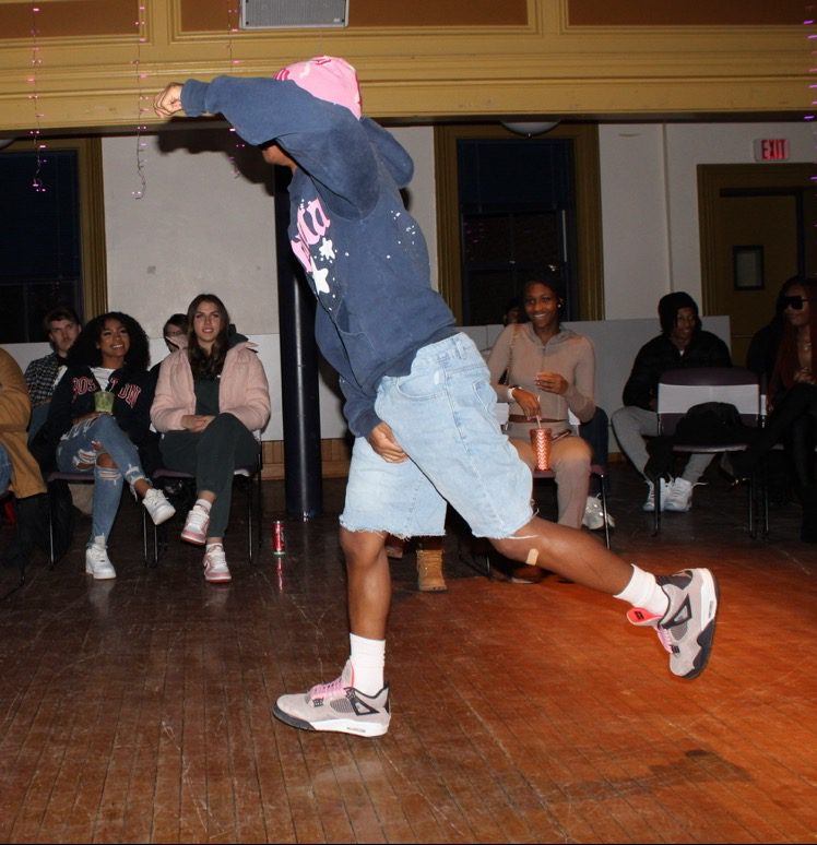 A young man dressed in a pink beanie, grey sweater, jorts and grey sneakers dances in a room with an audience seated in a semicircle.