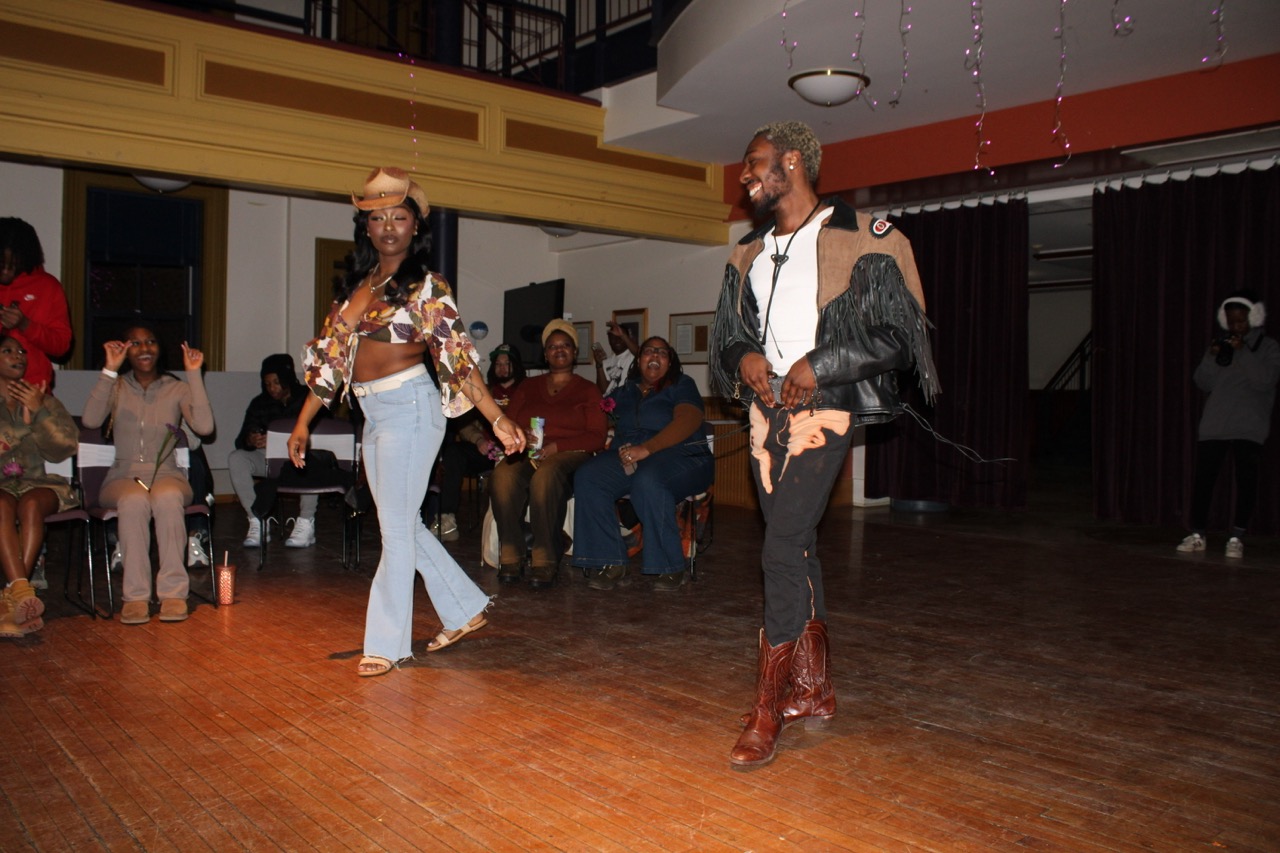 Two people walk on a wooden floor, with an audience seated in the background. The individual on the left is wearing a cowboy hat, cow print crop top, jeans and sandals. The individual on the right is wearing a white t-shirt, bolo tie, fringed jacket, jeans and dark brown cowboy boots.