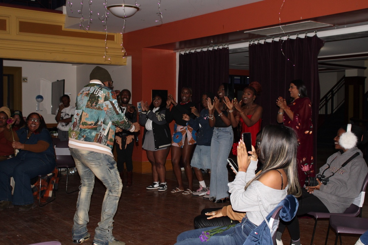 An individual with their back to the camera poses in a room surrounded by a clapping audience. The individual is dressed in a multicolored varsity jacket, jeans and grey sneakers.