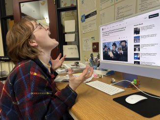 A young woman wearing a plaid shirt sits at a desktop computer displaying a news headline about the groundhog’s prediction for six more weeks of winter. The woman leans her head back in anguish and throws her hands in the air.