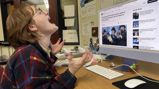 A young woman wearing a plaid shirt sits at a desktop computer displaying a news headline about the groundhog’s prediction for six more weeks of winter. The woman leans her head back in anguish and throws her hands in the air.