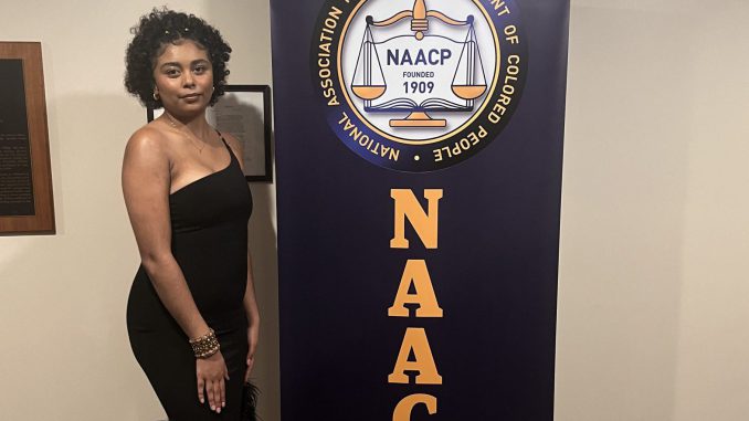 A young Black person stands in a black dress next to a blue drop down banner that says "NAACP" in yellow letters.