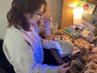 A young adult sits at a desk with one hand under a white UV lamp and the other holding nail polish. Beside the UV lamp is a cluster of cords.