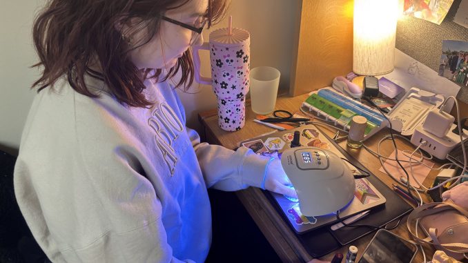 A young adult sits at a desk with one hand under a white UV lamp and the other holding nail polish. Beside the UV lamp is a cluster of cords.