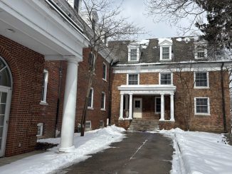 A shoveled path leading to a brick building with white pillars. The ground in front of it is covered in snow beside the pavement.