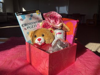 A red square box with hearts sits on a bed of pink tissue paper. Inside the box are a teddy bear, fake flowers, a canned beverage, a book of crossword puzzles and some assorted chocolates.