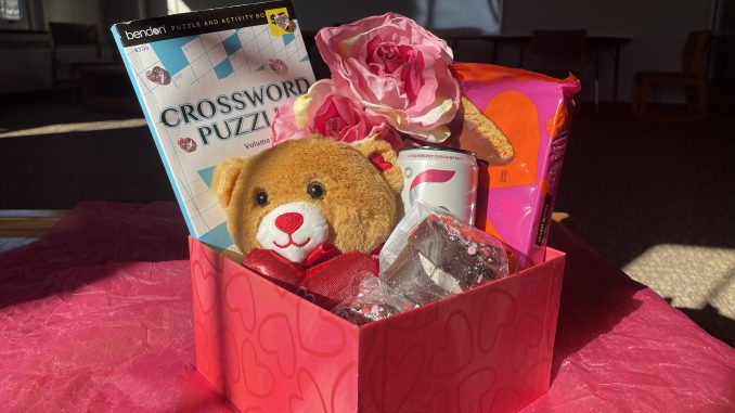 A red square box with hearts sits on a bed of pink tissue paper. Inside the box are a teddy bear, fake flowers, a canned beverage, a book of crossword puzzles and some assorted chocolates.