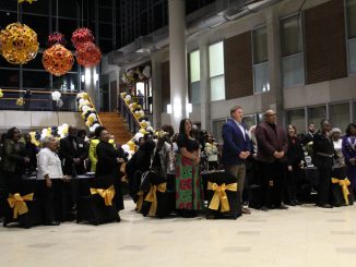 A group of people stand beside black chairs with yellow bows on the back in a large hall decorated with the colors black, yellow and white. Each guest is dressed in formal attire, and are looking toward something in the distance.