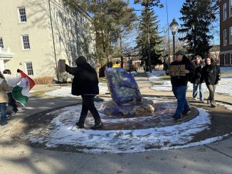 Seven young adults walk around a large purple and gold painted rock outside. Surrounding the rock there is snow on the ground. Two of the people hold a Mexican flag between them, and two others hold cardboard signs; one of the signs reads “F— I.C.E.”