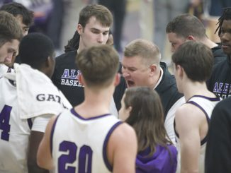 A circle of men in purple and white basketball jerseys surrounds an older man who is seen yelling. Behind the man are multiple people in black shirts reading “Albion Basketball”.