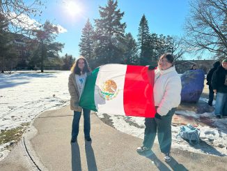 Two students stand holding the Mexican flag on a sidewalk, surrounded by snow. Other students stand in the background around a rock that is painted purple.