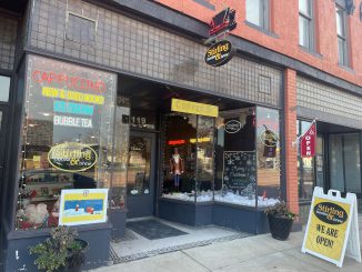 A storefront with tall glass windows sporting lots of signage with a dark trim around the bottom. A yellow sign is propped up on the sidewalk next to the store, reading “Stirling books & brew, we are open!”