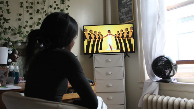 A girl sits in a brightly lit room in a chair beside a desk with her hair pulled back in a clip. She sits facing a tv where a yellow screen with animated characters stand around boiling pots and miscellaneous cooking supplies.