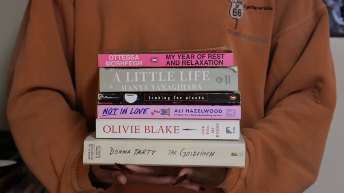 Two hands hold a stack of six different books, differing in color and thickness, with their spines facing the camera. Uhhh I can probably put more here but would like a little help.