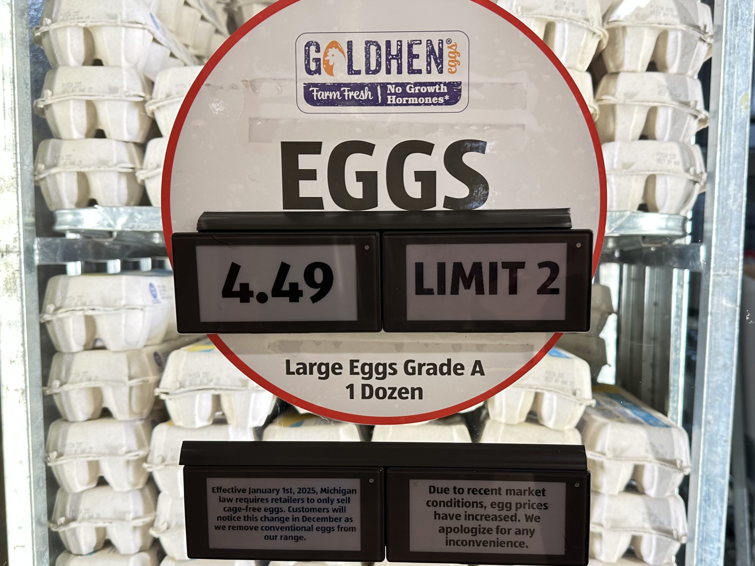 A sign reading “Eggs, 4.49, Limit 2” is displayed on a glass door to a cooler full of egg cartons. Two smaller signs below describe prices rising due to Michigan’s cage-free egg law and recent market conditions.