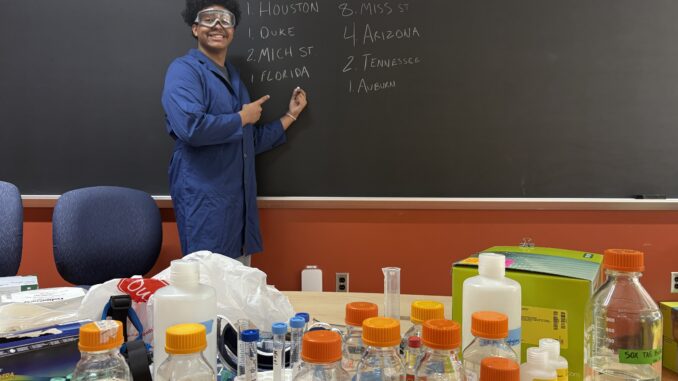 A young man in a blue lab coat and safety goggles stands in front of a blackboard, smiling and pointing at a list of college basketball teams written in chalk. In the foreground is a table filled with various labeled lab bottles, test tubes and scientific equipment.