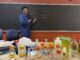 A young man in a blue lab coat and safety goggles stands in front of a blackboard, smiling and pointing at a list of college basketball teams written in chalk. In the foreground is a table filled with various labeled lab bottles, test tubes and scientific equipment.