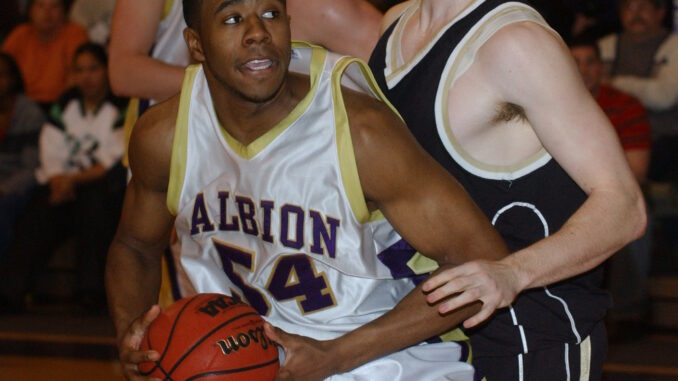 Basketball players in intense game action; one in white jersey with "Albion" attempts a shot while being defended by a player in black. Crowd watches intently.