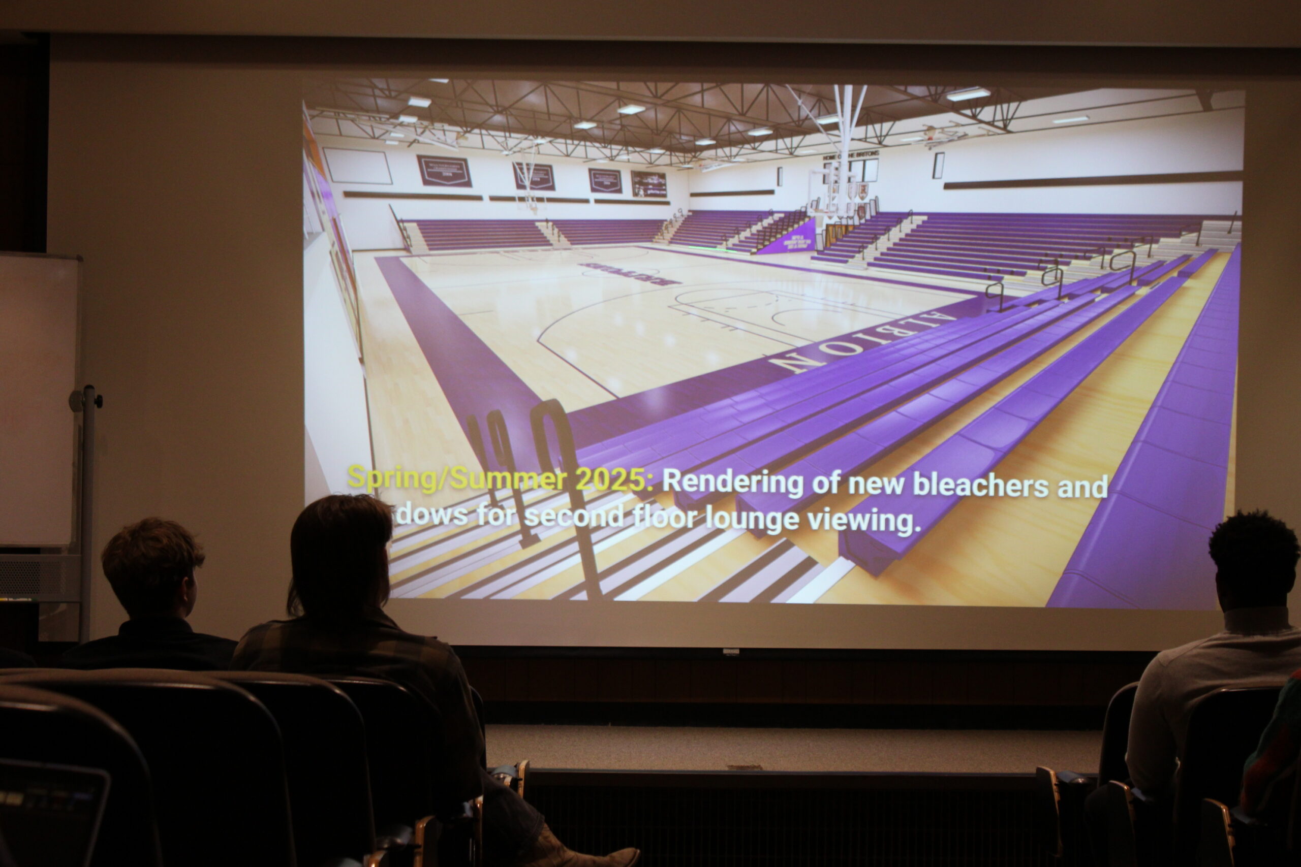 An audience viewing a projected rendering of a basketball court with purple bleachers surrounding three of the four sides. The bottom left has text that reads, “Spring/Summer 2025: Rendering of new bleachers and windows for second lounge floor viewing.”