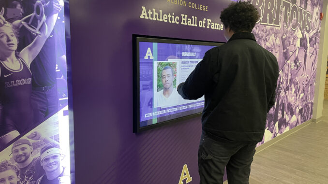 A young adult interacts with a digital display with the title “Albion College’s Athletic Hall of Fame.” Behind the display is a wall featuring images of Albion sports, and the photos are covered in a purple filter.