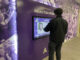 A young adult interacts with a digital display with the title “Albion College’s Athletic Hall of Fame.” Behind the display is a wall featuring images of Albion sports, and the photos are covered in a purple filter.