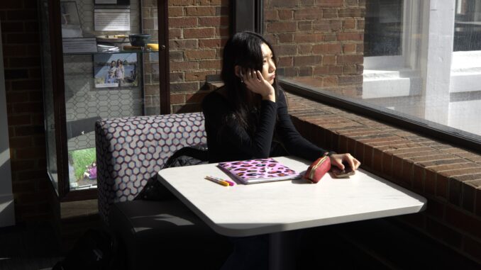 A young woman sits on a purple patterned chair with a gray table in front of her with a folder, pens and a red purse splayed on top. She rests her head on her right palm as she looks out a window with a brick ledge below it.