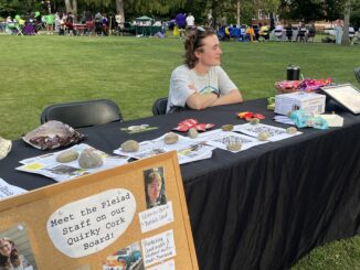 A young white woman with short brown hair sits behind a table covered with a black table cloth and several papers and packages of colorful candy. The woman has her arms crossed sitting on the table, and looks to the right, smiling. Behind her are several other tables in the distance, and in the foreground there is a cork board with photos of other people and a note reading “Meet the Pleiad Staff on our Quirky Cork Board!”