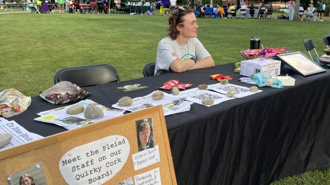 A young white woman with short brown hair sits behind a table covered with a black table cloth and several papers and packages of colorful candy. The woman has her arms crossed sitting on the table, and looks to the right, smiling. Behind her are several other tables in the distance, and in the foreground there is a cork board with photos of other people and a note reading “Meet the Pleiad Staff on our Quirky Cork Board!”