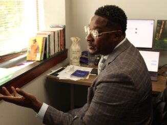 A middle-aged Black man in a gray suit gestures with an open hand toward a window while speaking. Sunlight illuminates his glasses as well as the desk behind him, on which stand three computer monitors, several papers, and books leaning against the window sill.