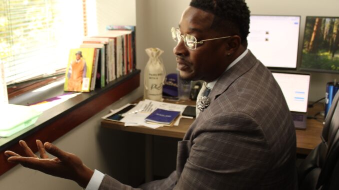 A middle-aged Black man in a gray suit gestures with an open hand toward a window while speaking. Sunlight illuminates his glasses as well as the desk behind him, on which stand three computer monitors, several papers, and books leaning against the window sill.