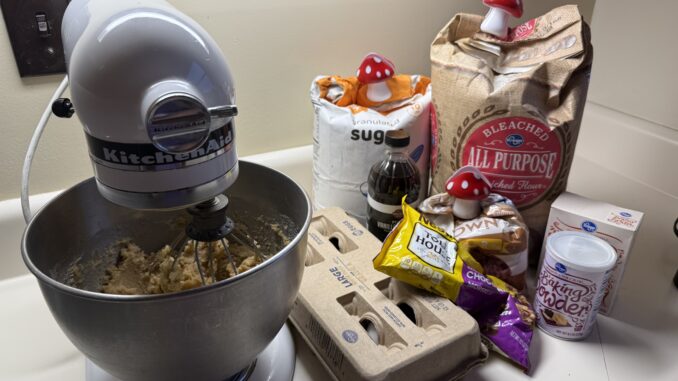 A KitchenAid mixer stands on the left with cookie batter inside. To the right are ingredients for chocolate chip cookies: eggs, vanilla, sugar, flour, brown sugar, chocolate chips, baking powder and baking soda.